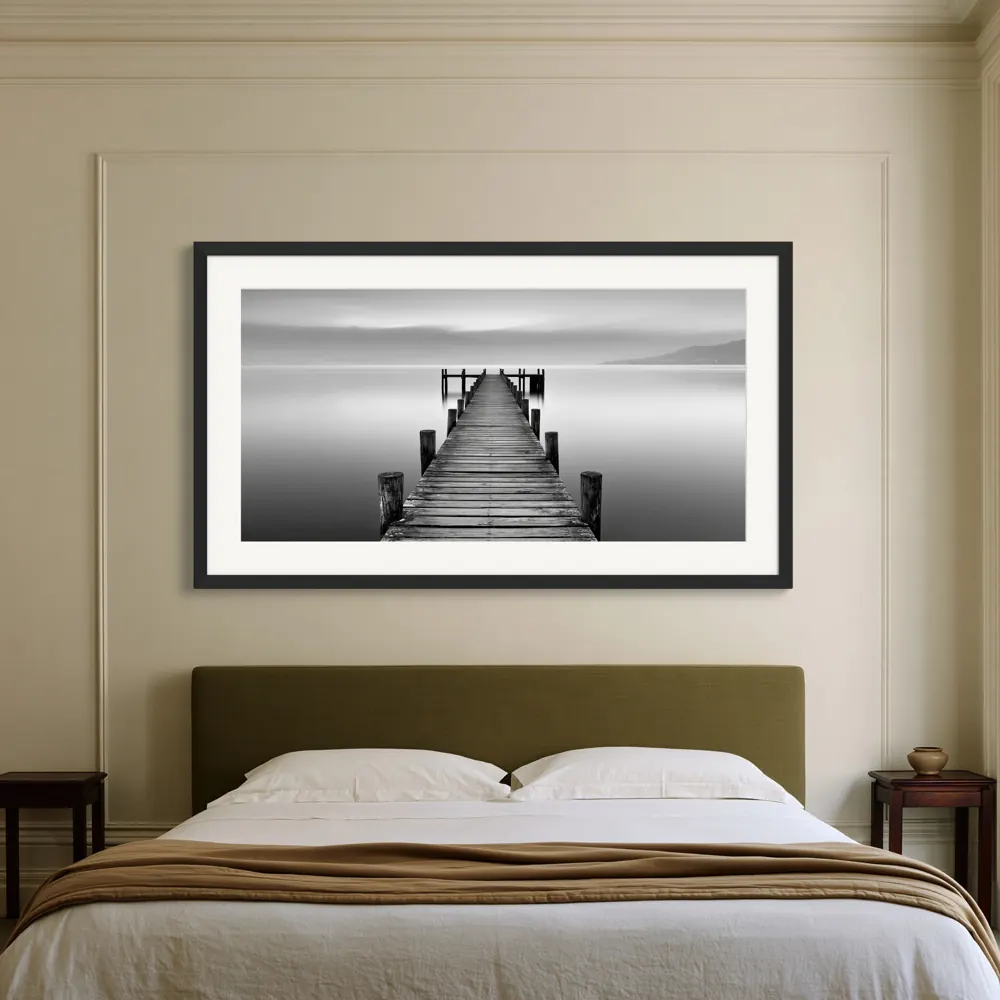 Black and white photograph of a wooden pier in a black frame with a wide white mat above a bed with an olive headboard