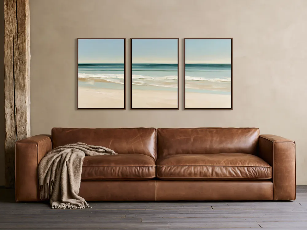 Three framed beach paintings in a row above a tan leather sofa