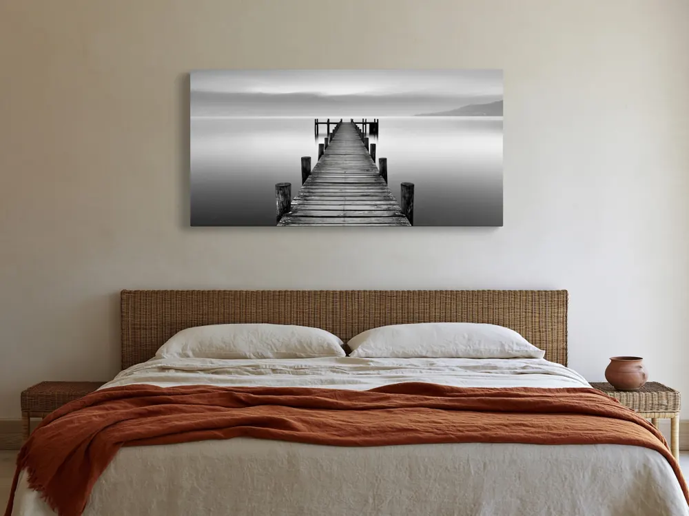 A black-and-white lake pier photograph as a stretched canvas in the Limewash bedroom with cane headboard