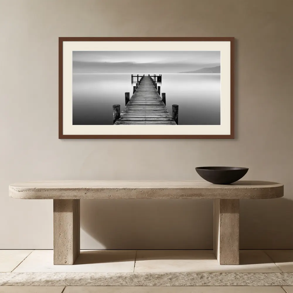 A black-and-white lake pier photograph as a walnut framed print with a warm-white mat in the Travertine entry console